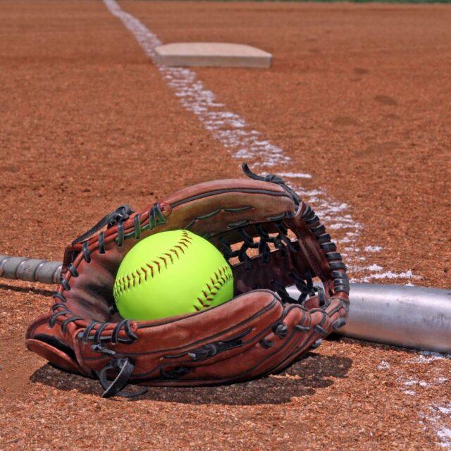 A softball resting inside a glove on a baseball field near home plate.
