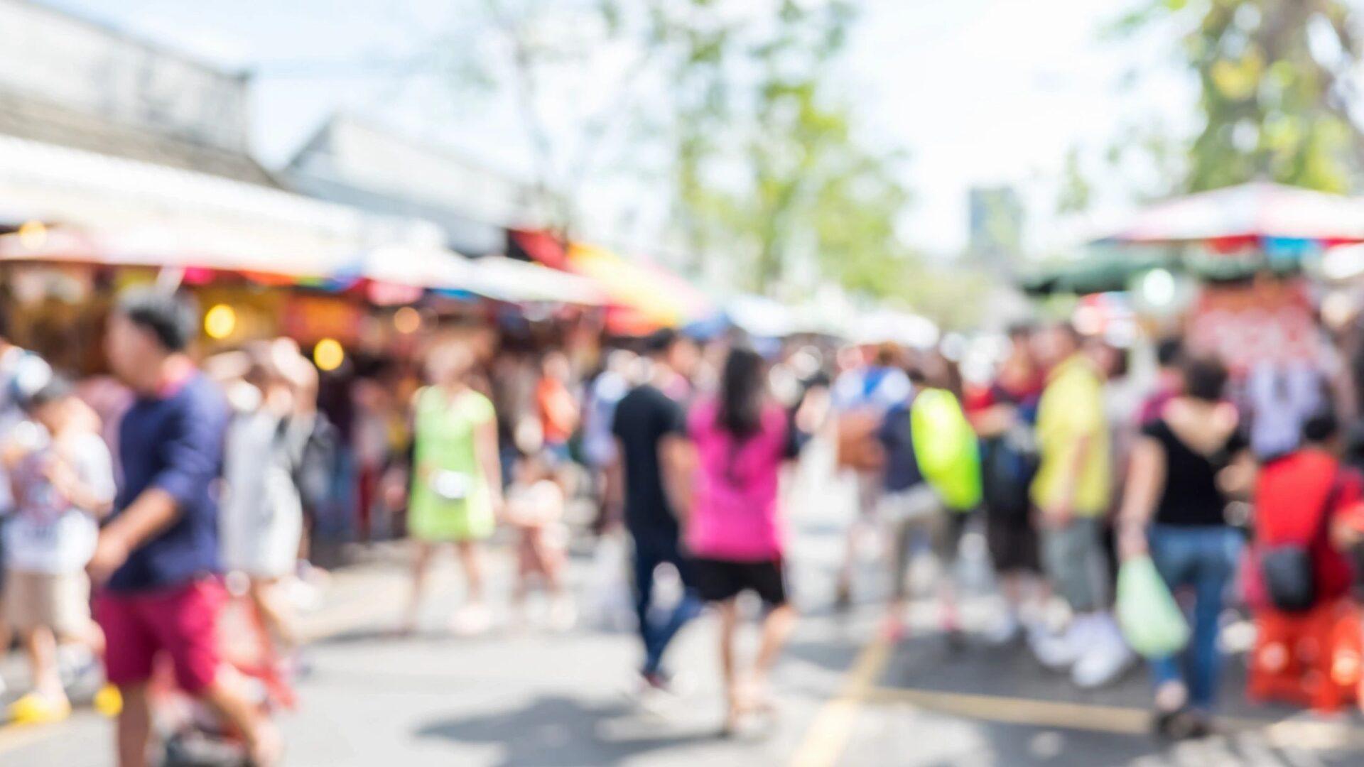 Blurred image of people walking at an outdoor market or fair on a sunny day.
