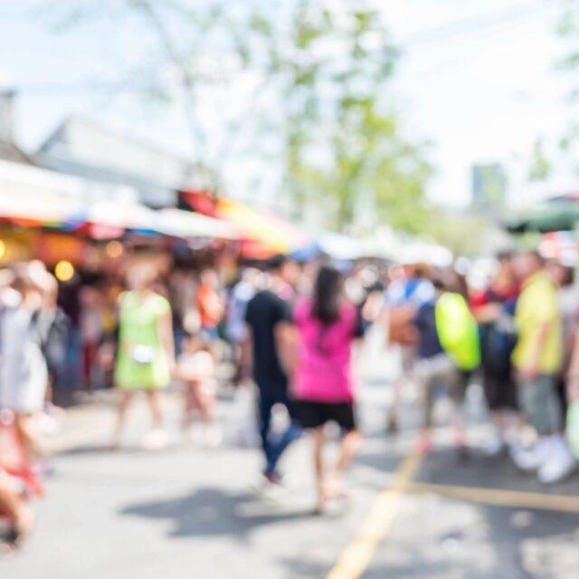 Blurred image of people walking at an outdoor market or fair on a sunny day.