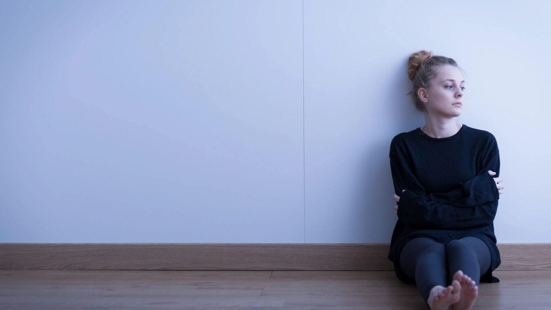Woman sitting alone against a wall, looking contemplative.