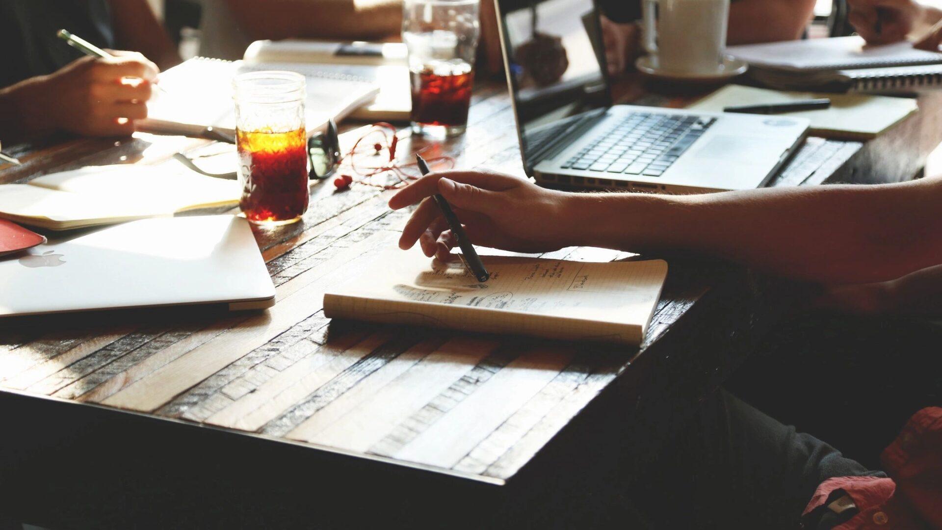 Person writing notes with a laptop and drinks on the table.