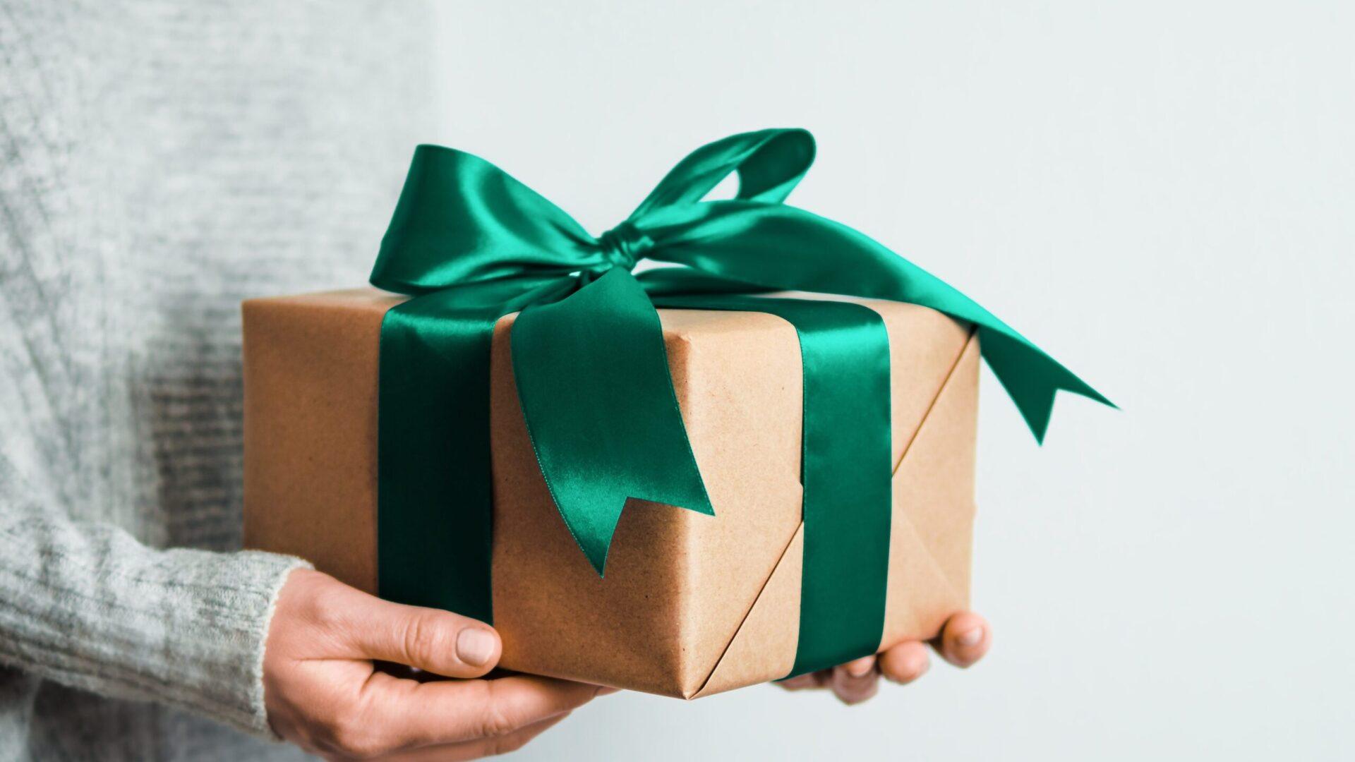 A hand holding a gift wrapped in brown paper with a green ribbon.